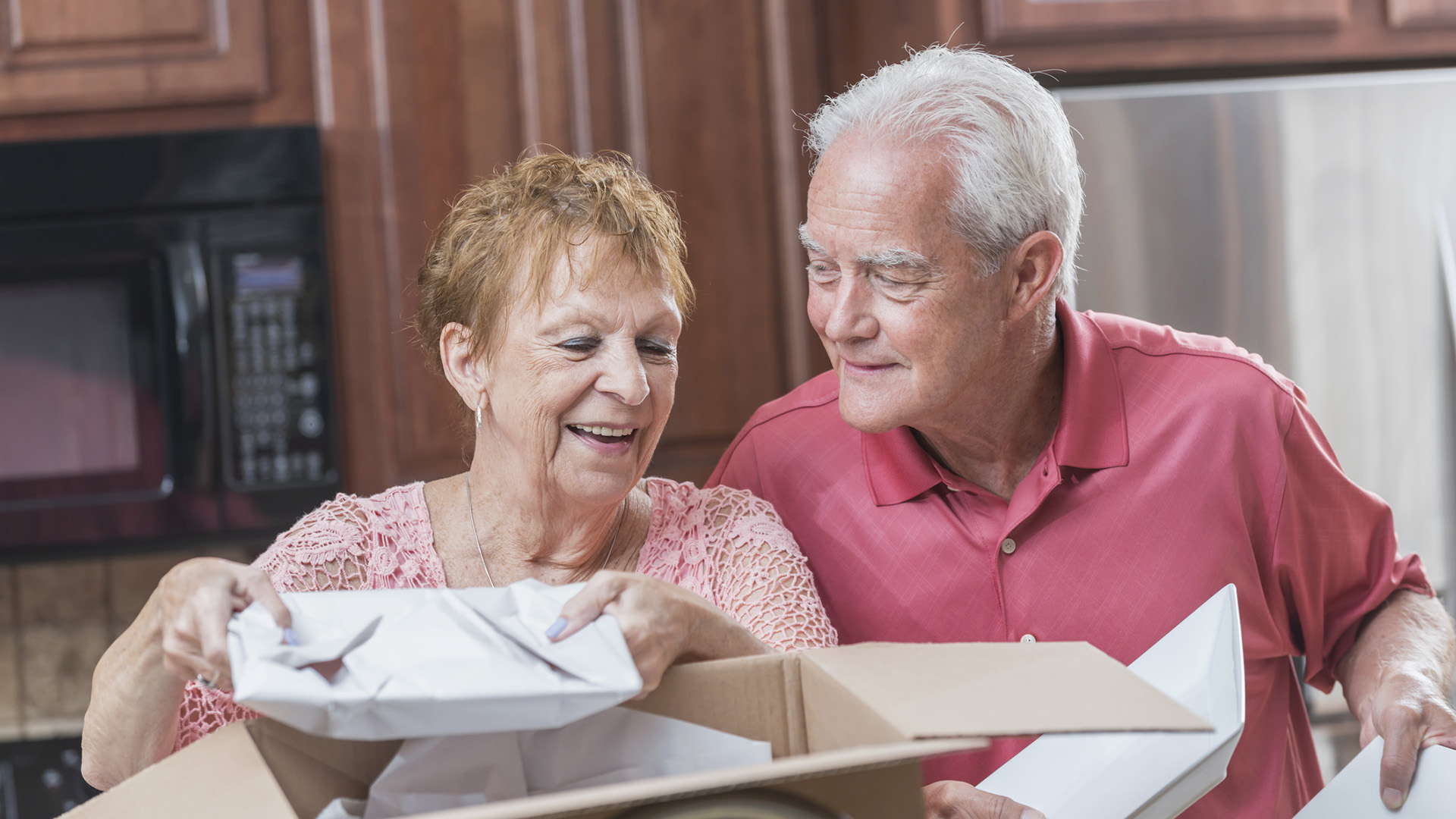 A senior couple moving house. They are packing or unpacking a cardboard box with dishes in the kitchen. They are smiling and talking as they lift the plates.